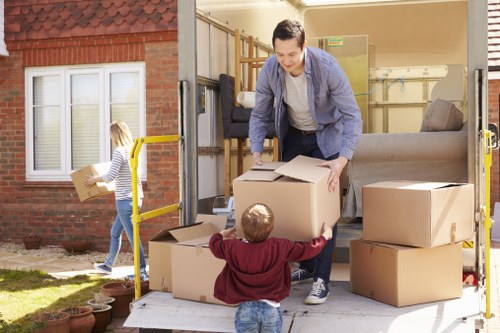 A male mover in casual clothing is loading cardboard moving boxes onto a van parked in a residential driveway, with several boxes stacked inside the van and on the ground around it. A young girl with blonde hair, wearing a striped shirt and jeans, is carrying a box towards the back of the van, while a small boy dressed in a burgundy sweatshirt reaches up, possibly to help or receive a box. The background features a red-brick house with white-framed windows and potted plants along the base, indicating a typical neighbourhood setting in Brent or nearby area. The scene captures the process of house removals, showing the careful handling of household items like cardboard boxes on a sunny day with natural light illuminating the area. The interior of the van suggests it is being filled with furniture and personal belongings in preparation for a move, consistent with professional removals services offered by Brent Man with Van.