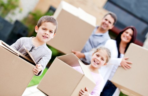 A family of four, including a young boy and girl, is standing outdoors in a residential area, holding various cardboard boxes as part of a house move. The children are smiling and appear engaged in the packing process. The boy in the foreground is wearing a light grey long-sleeved shirt and is holding a large cardboard box. Behind him, a woman with dark hair, dressed in a white top, and a man with glasses and dark hair, dressed in a light blue shirt, are also holding boxes and smiling. The background shows a modern house with large windows and a garden with some greenery, indicating a suburban setting. The scene is well-lit with natural daylight, emphasizing a friendly and organized moving effort. This image relates to house removals and professional moving services, exemplifying family participation in a local move, possibly in Brent or nearby areas within the postcode region of SW or NW London.