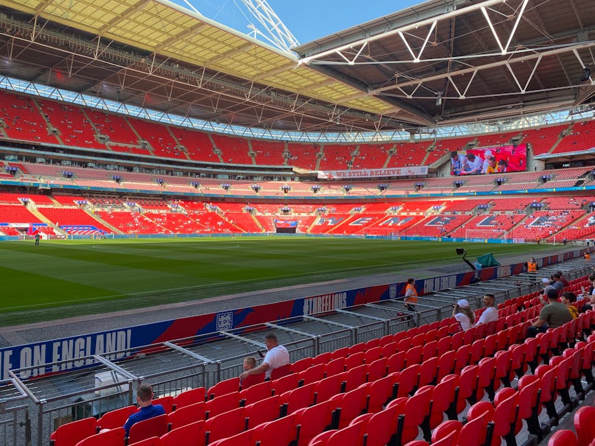 Inside Wembley Stadium, the image shows a large, modern sports arena with red tiered seating surrounding a well-maintained grass pitch. The stands are mostly empty, and the word 'WEMBLEY' is visible s